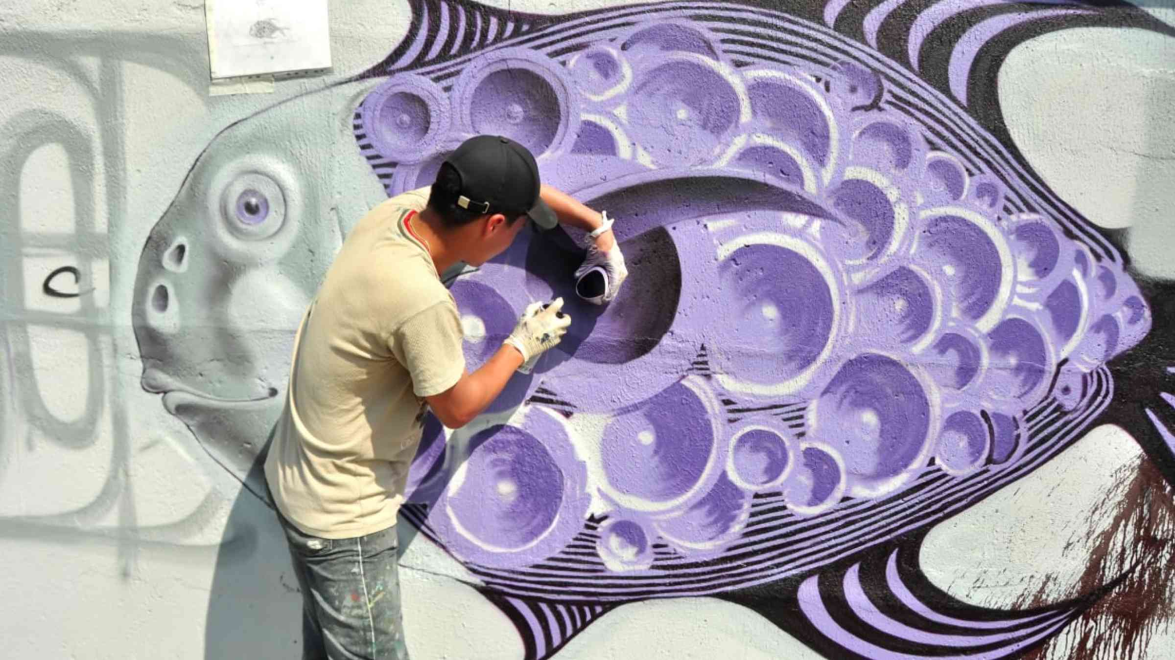 Graffiti artist painting a  purple fish to a wall.