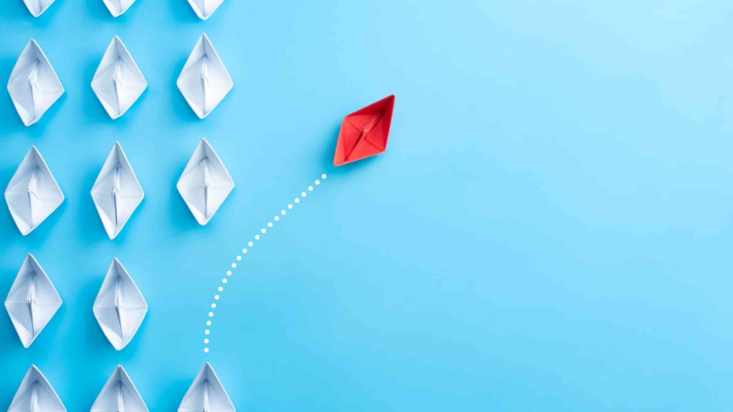 Group of white paper ship in one direction and one red paper ship pointing in different way on blue background.