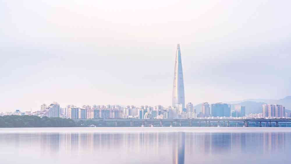 The skyline of a large city with a modern skyscraper soaring above the other buildings, reflected in the water. The photo has a purple hue and was likely taken at dusk.
